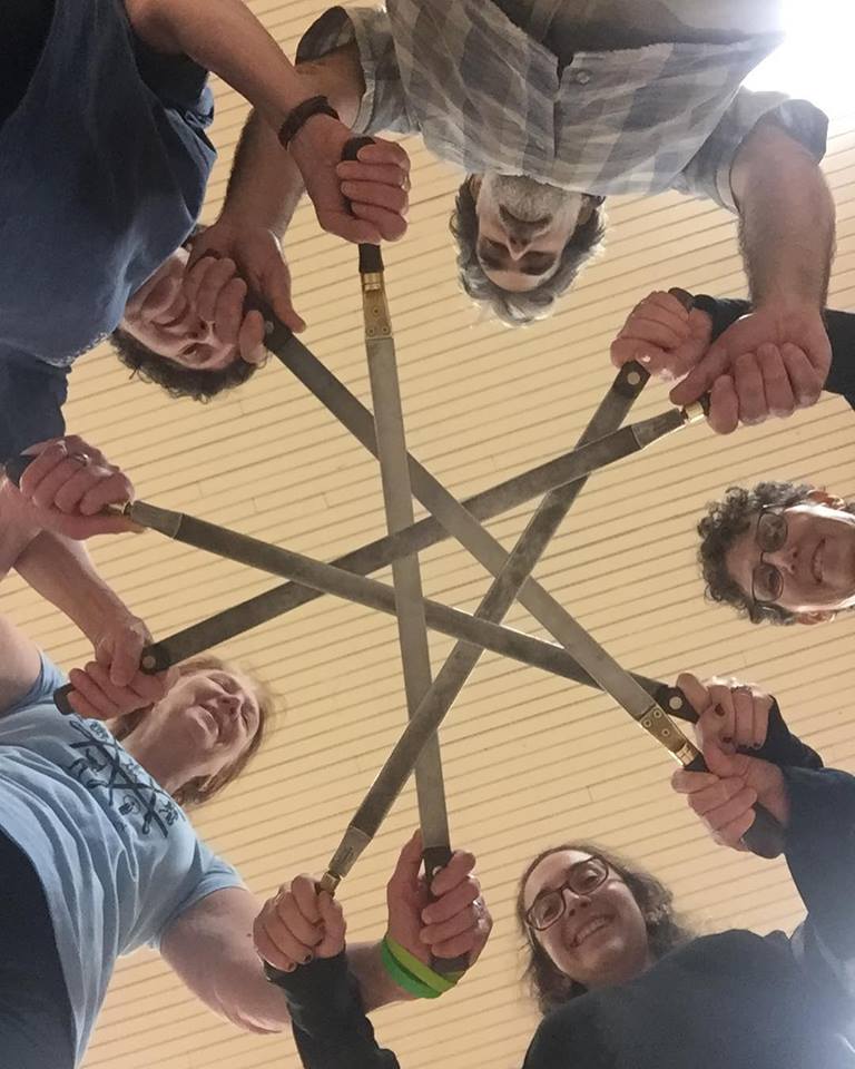 5 people stand in a circle holding swords to form the shape of a star
