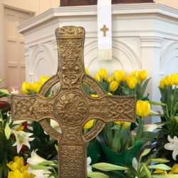 A celtic cross stands in front of lilies and daffodils with the pulpit behind