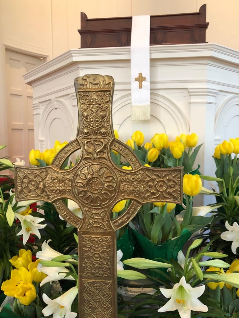 A celtic cross stands in front of lilies and daffodils with the pulpit behind